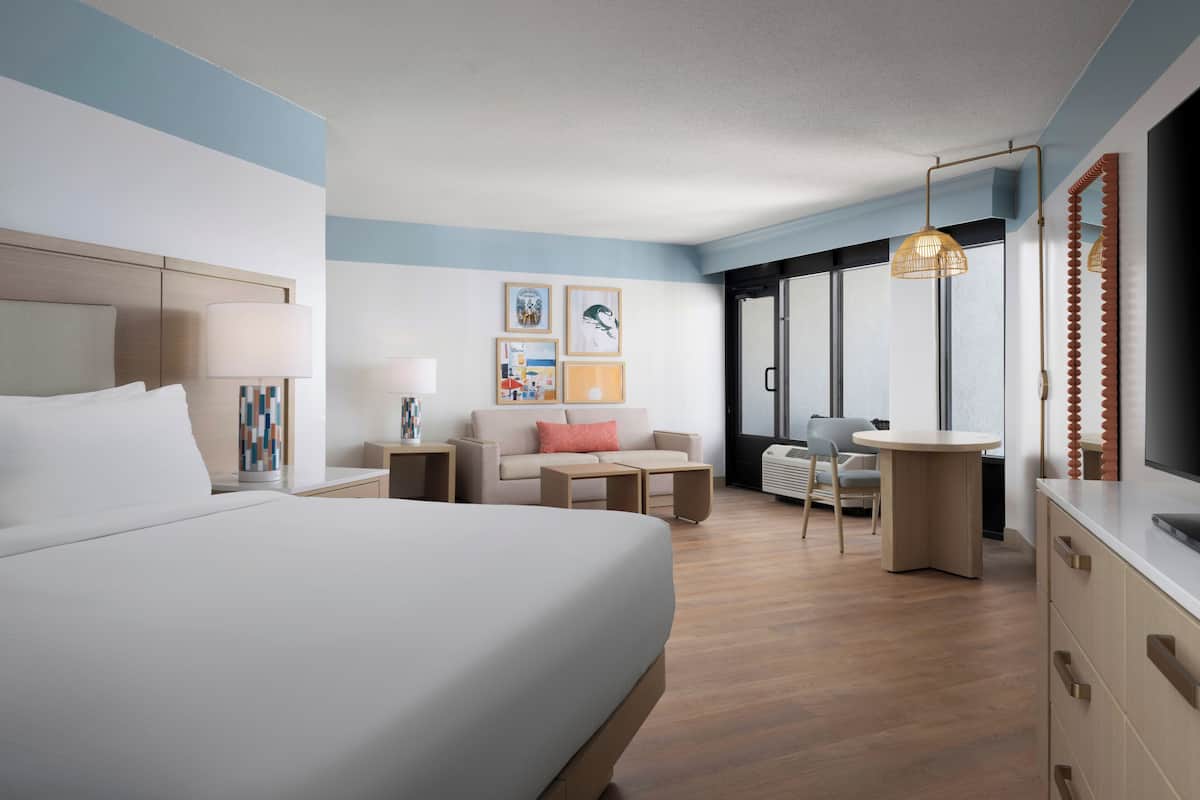 A modern hotel room with a large bed, a sofa, a small table, and a desk. There are framed pictures on the wall, a window with a view, and a ceiling light.