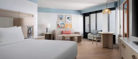 A modern hotel room with a large bed, a sofa, a small table, and a desk. There are framed pictures on the wall, a window with a view, and a ceiling light.