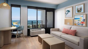 A modern living room with a sofa, a desk, and a chair, a large window with a view of the sea, and artwork on the wall. - The Ellie Beach Resort Myrtle Beach, Tapestry By Hilton (Myrtle Beach)