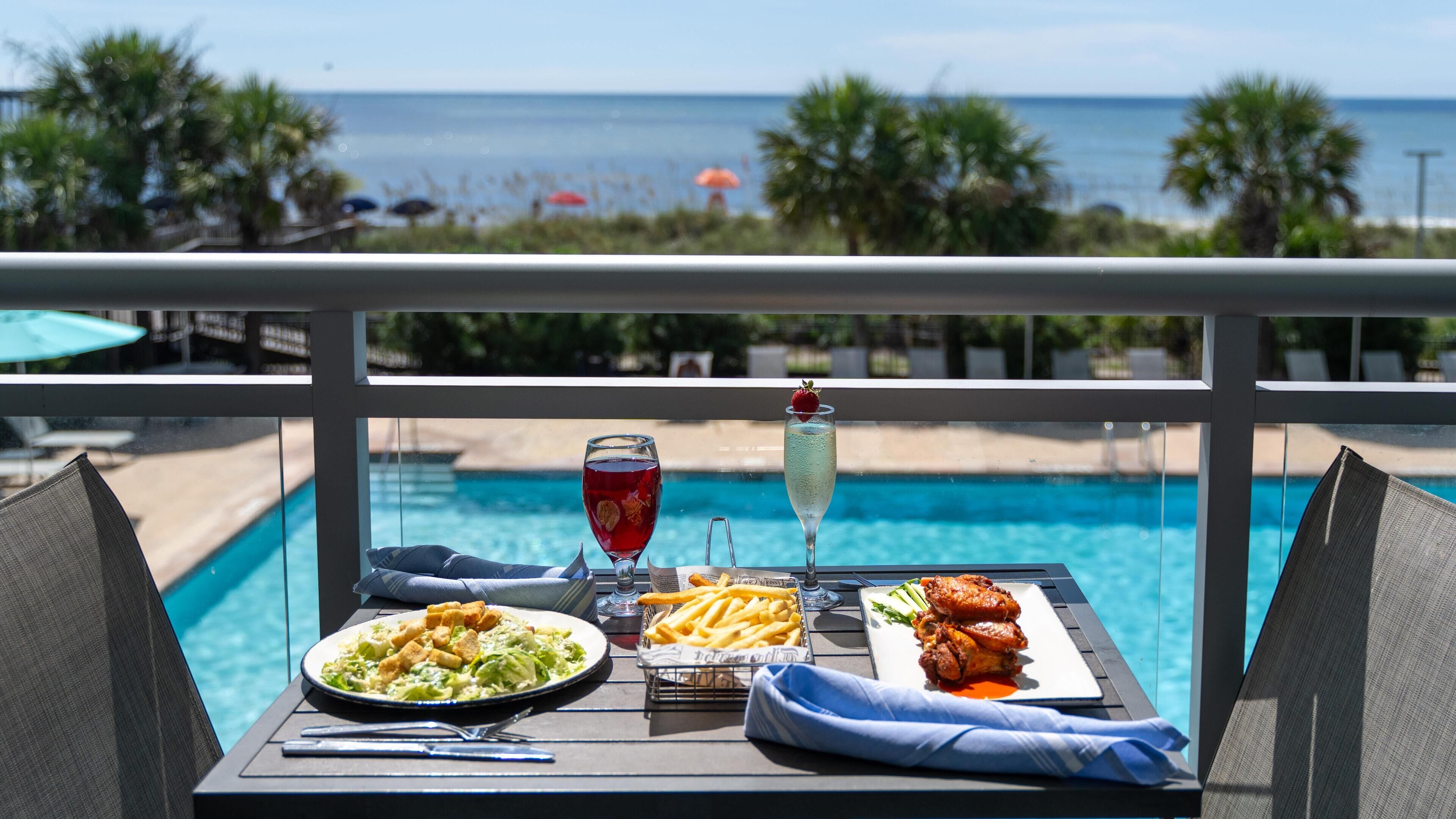 A dining table with a view of a pool and ocean, featuring a salad, fries, wings, and two drinks.