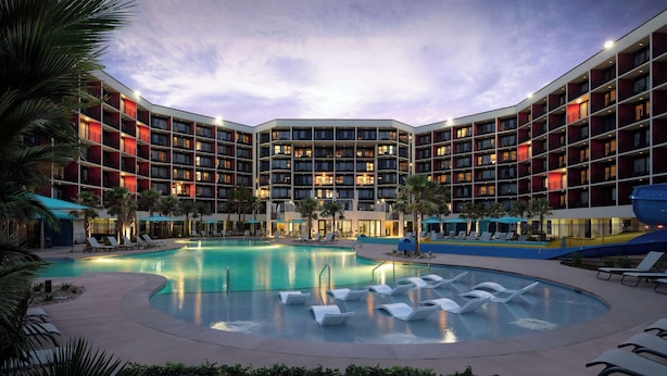 A hotel with a central pool surrounded by lounge chairs, palm trees, and a multi-story building with balconies. - The Ellie Beach Resort Myrtle Beach, Tapestry By Hilton (Myrtle Beach)