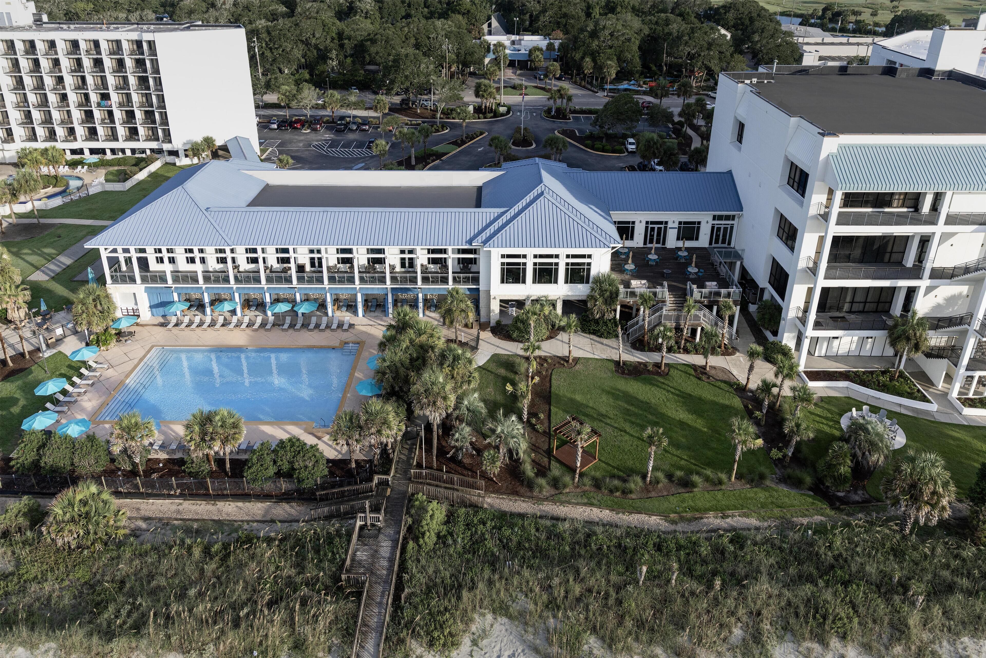 An aerial view of a hotel with a swimming pool, palm trees, and a parking area.