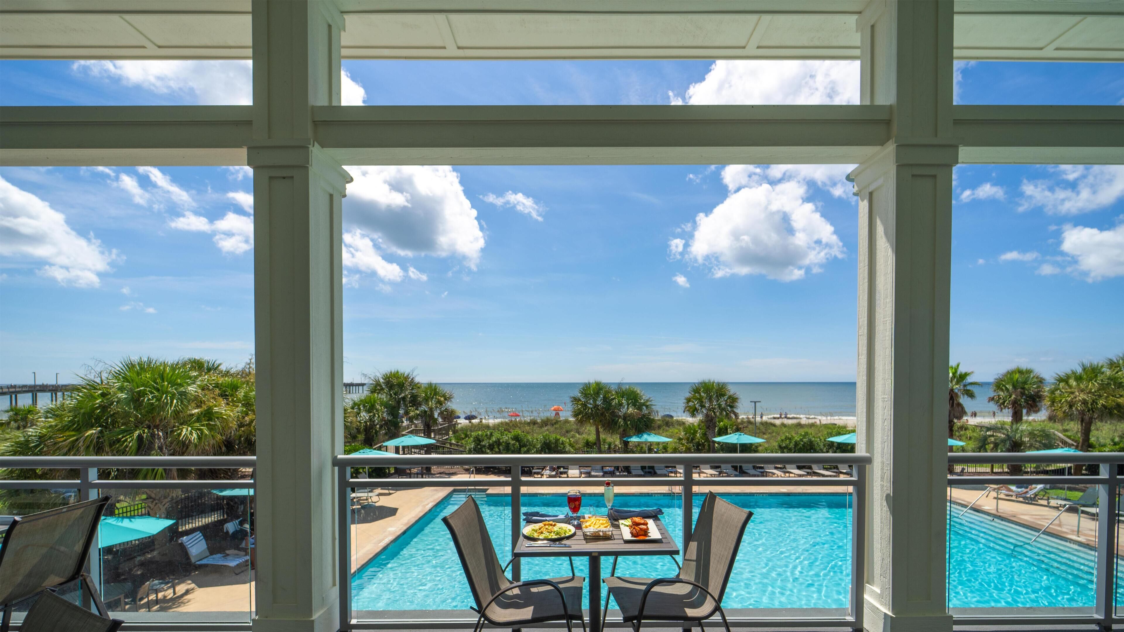 A poolside view with a table set for dining, overlooking a beach and the ocean.