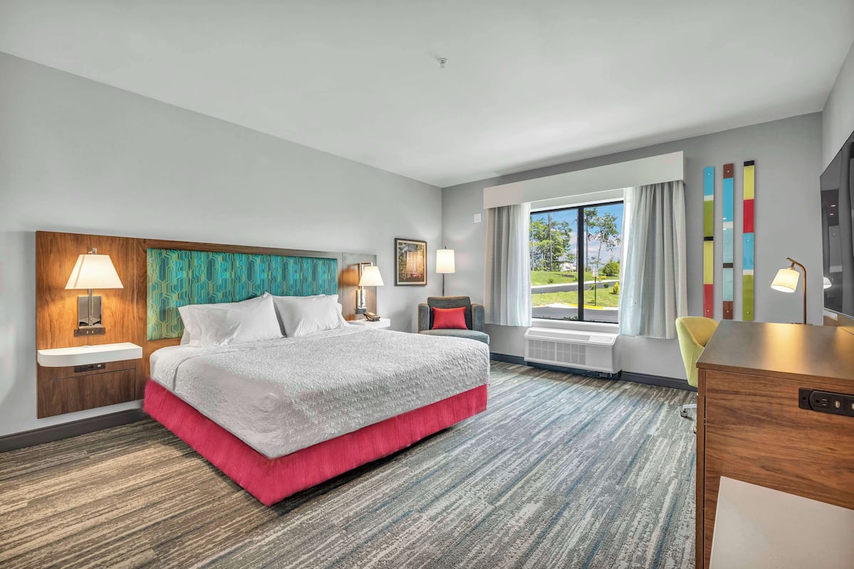 A hotel room with a large bed, two bedside tables with lamps, a desk, a chair, a window with curtains, and a view of the outdoors.