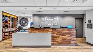A hotel reception area with a 'Welcome the Mother of States' sign, a wooden reception desk, and a variety of amenities on shelves.