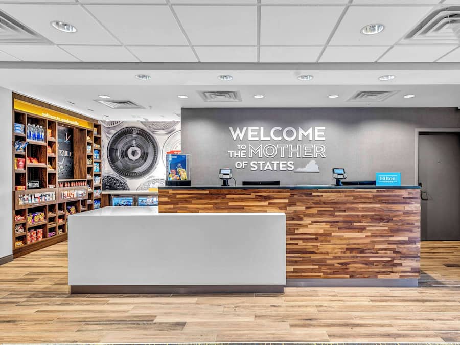 A hotel reception area with a 'Welcome the Mother of States' sign, a wooden reception desk, and a variety of amenities on shelves.