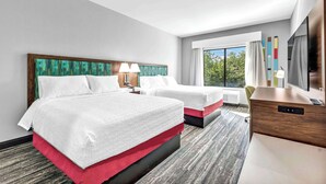 A hotel room with two beds, a desk, a chair, a TV, and a window with a view of trees.