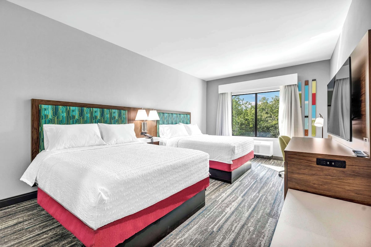 A hotel room with two beds, a desk, a chair, a TV, and a window with a view of trees.