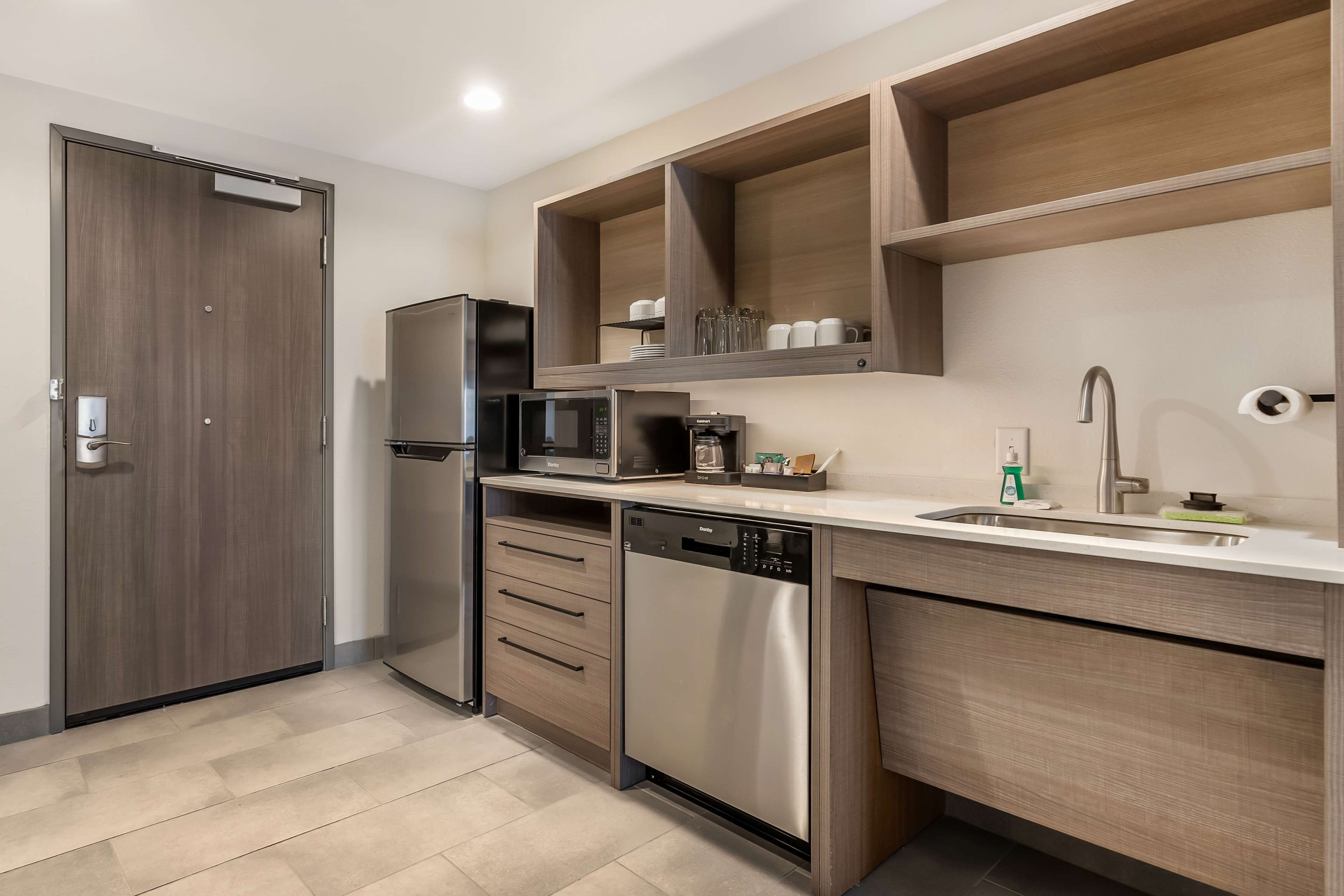 A modern kitchen with a built-in microwave, refrigerator, and dishwasher. There are wooden shelves above the counter and a sink with a faucet.