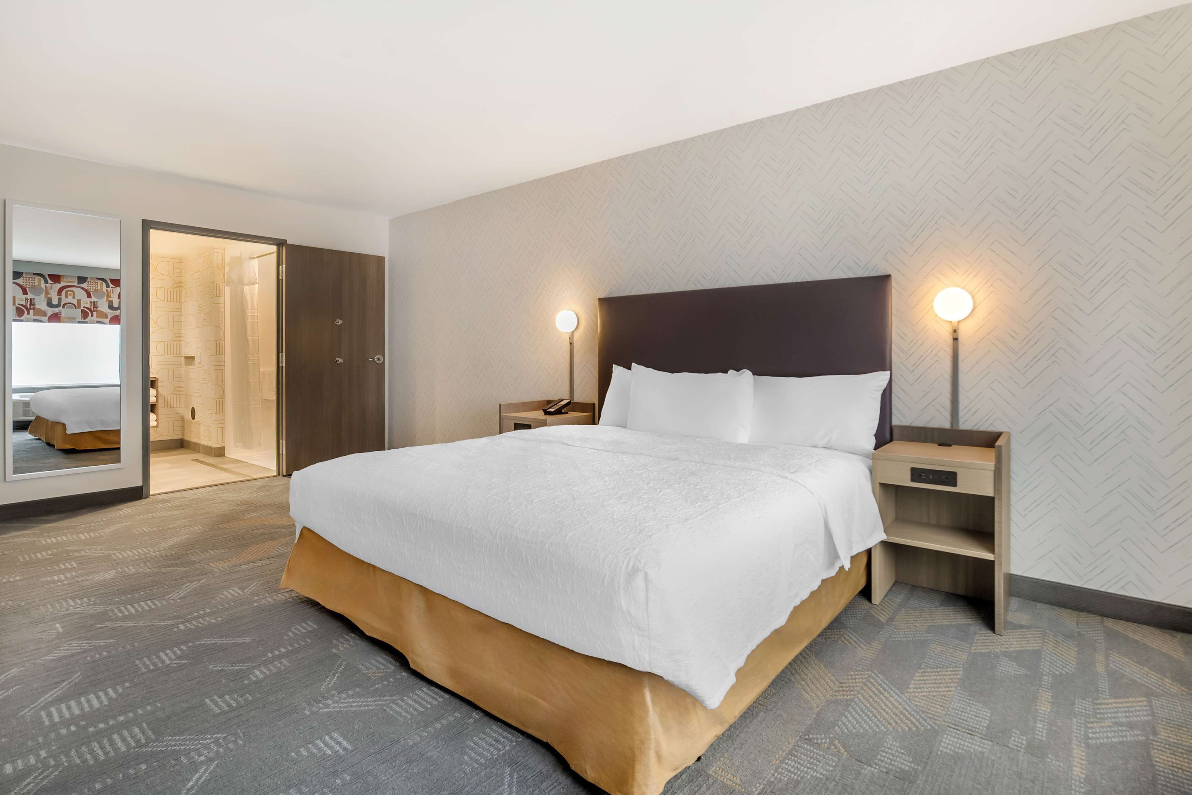 Hotellihuone, jossa on suuri sänky, kaksi yöpöytää, peili ja ovi, joka johtaa kylpyhuoneeseen.