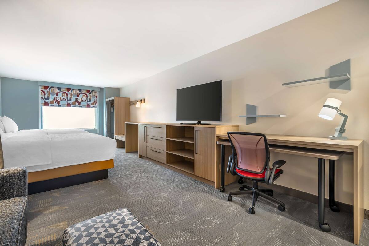 A hotel room with a bed, a desk, a chair, a TV, and a lamp.