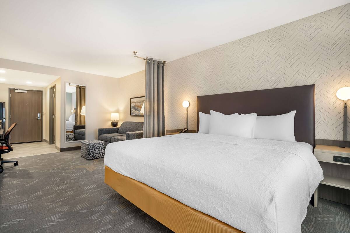 A hotel room with a large bed, a desk, a chair, a lamp, and a mirror.
