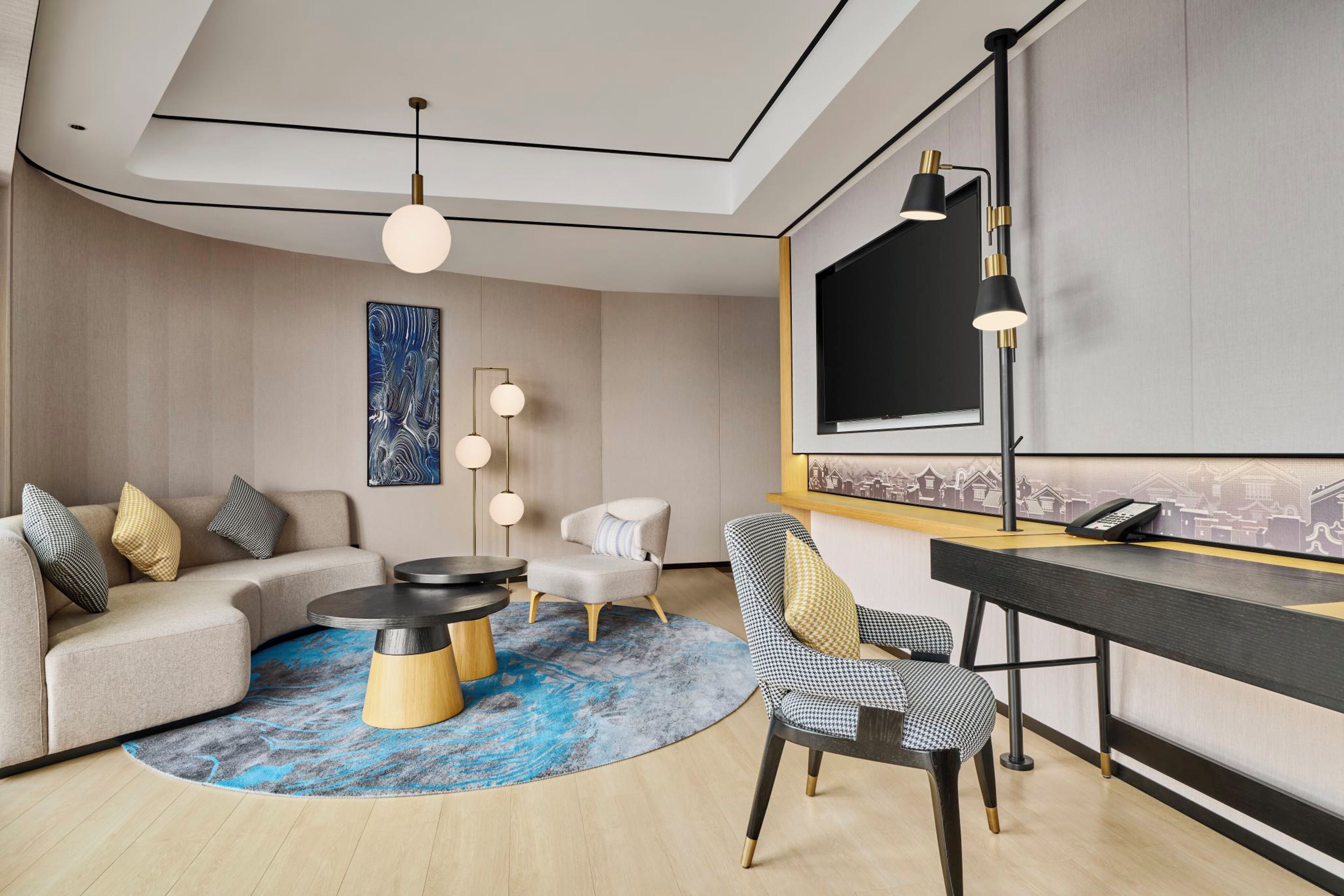 A modern hotel room with a curved ceiling, a round coffee table, a sofa, a desk, and a cityscape mural on the wall.