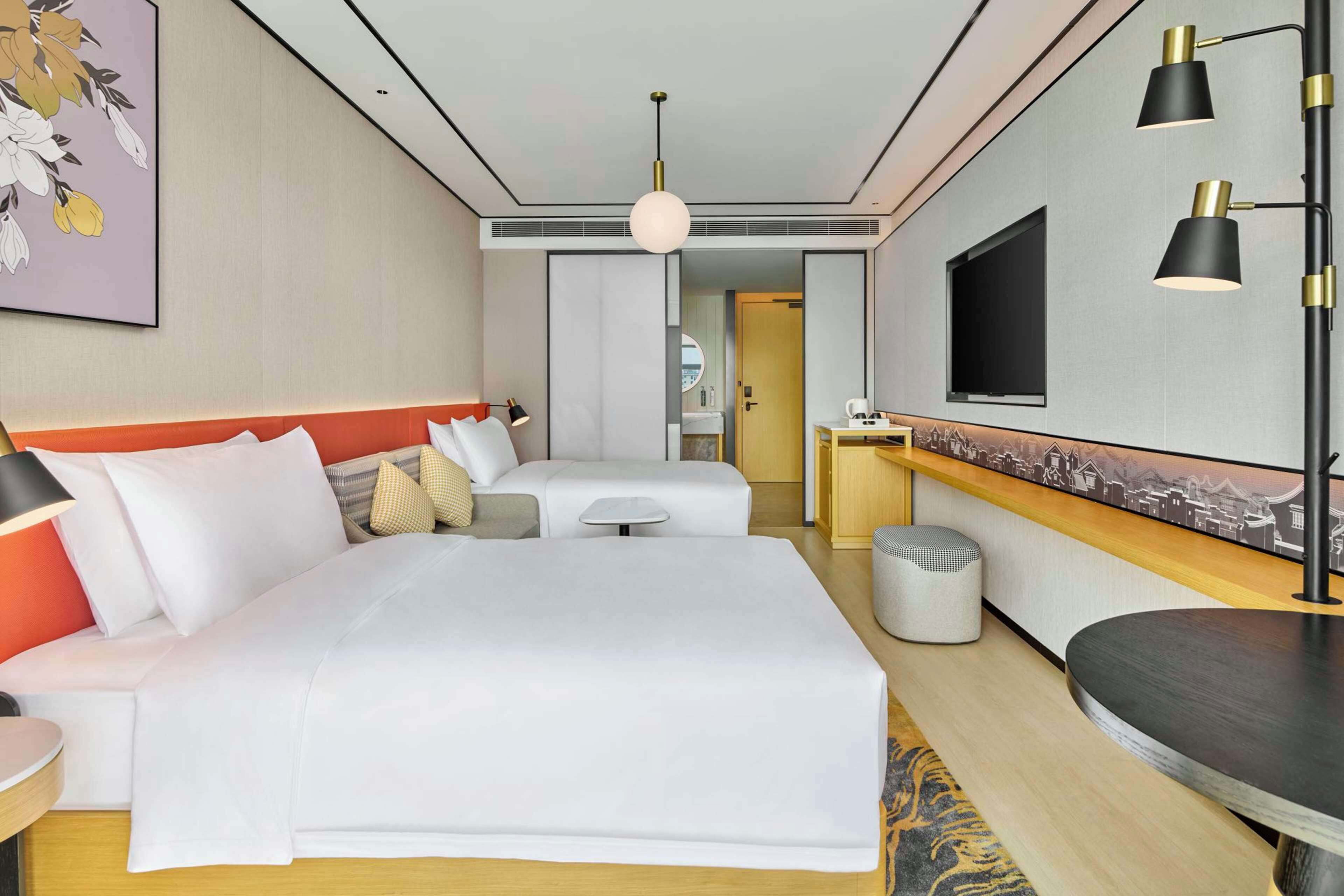 Ein Hotelzimmer mit einem großen Bett, einem Schreibtisch mit Stuhl, einem an der Wand montierten Fernseher und einem Gemälde über dem Bett.
