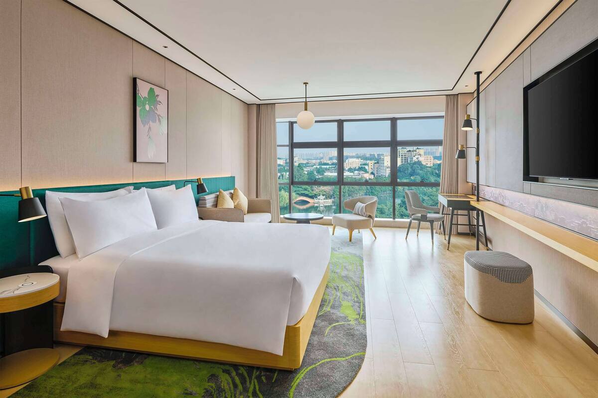 A modern hotel room with a large bed, a TV, a desk, and a view of the city.