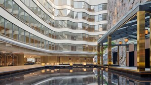 Ein modernes Hotelinneres mit mehreren Etagen, Glasgeländern und einem zentralen, spiegelnden Pool.