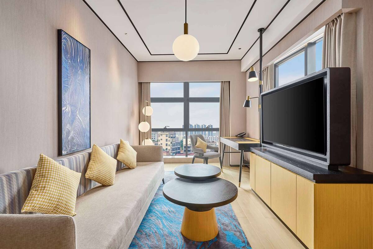 A modern hotel room with a sofa, a round table, a large TV, and a city view through the window.