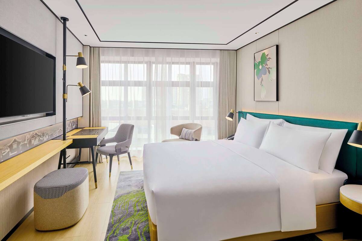 A hotel room with a large bed, a desk with a computer, a chair, a small ottoman, and a TV.
