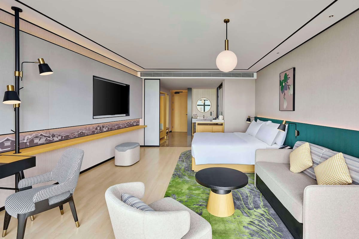 A modern hotel room with a bed, sofa, armchair, TV, and a desk with a chair.