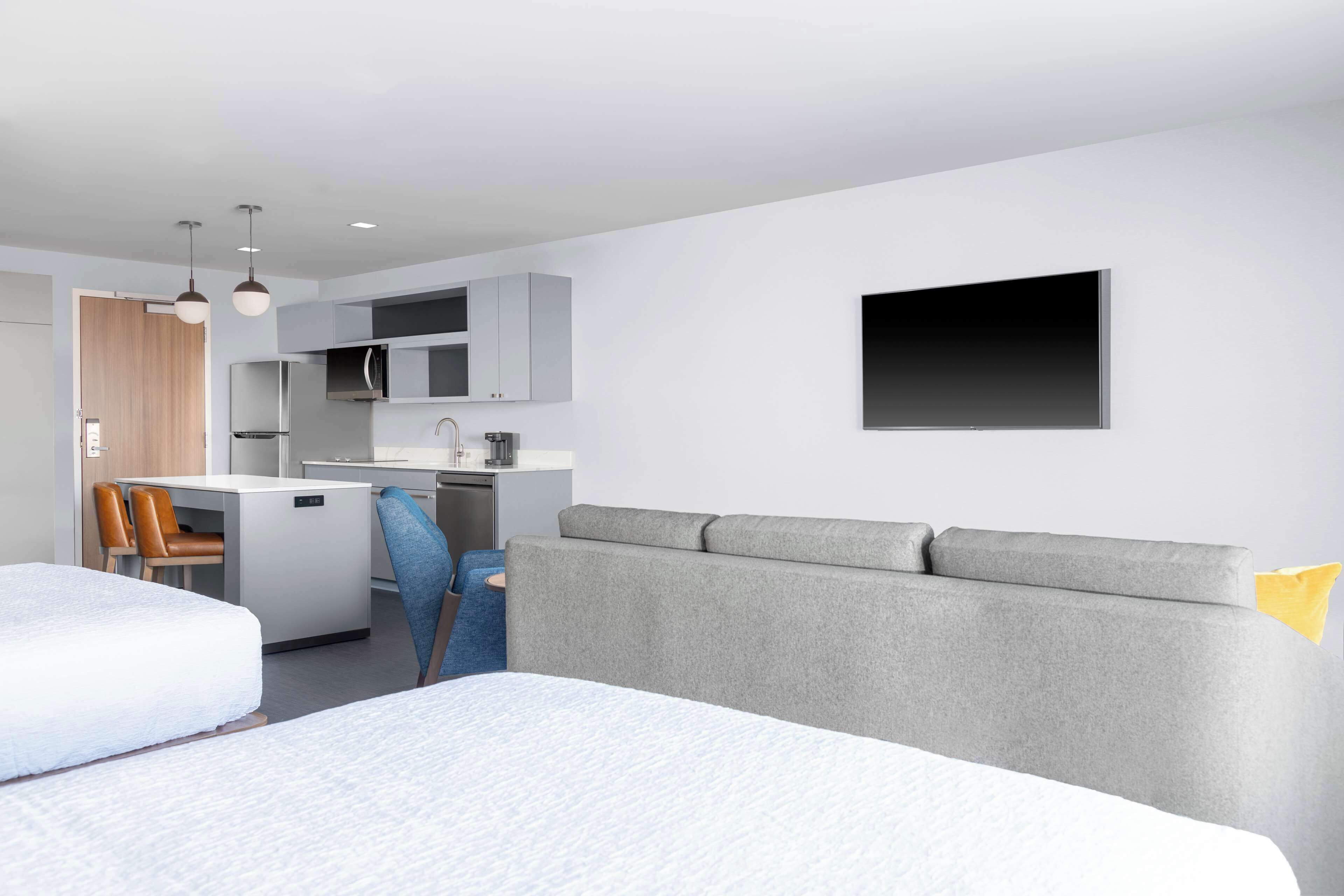 A modern hotel room with a kitchenette, a grey sofa, a bed, and a desk with a chair.