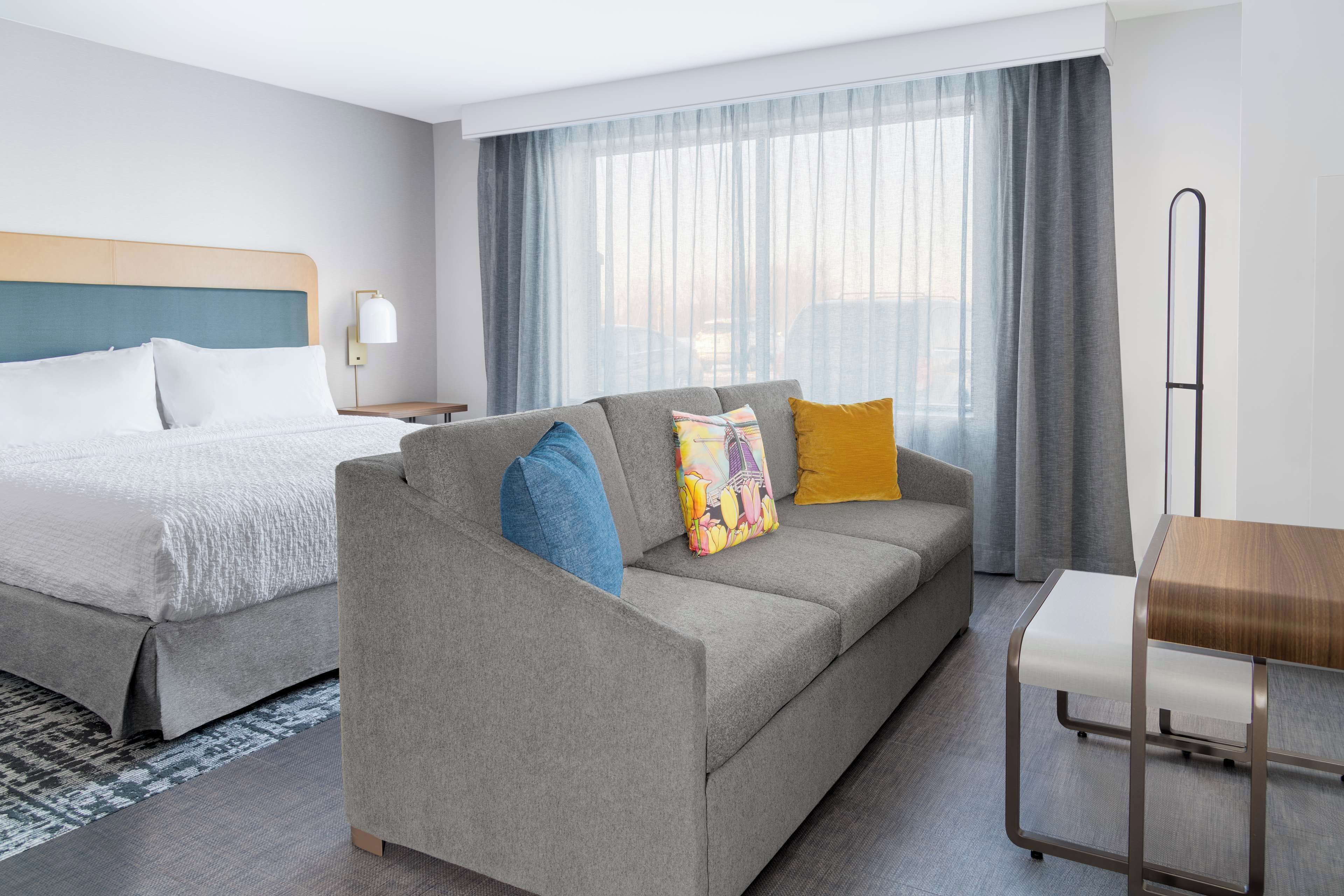 A hotel room with a bed, a sofa, a wooden table, and a large window with curtains.