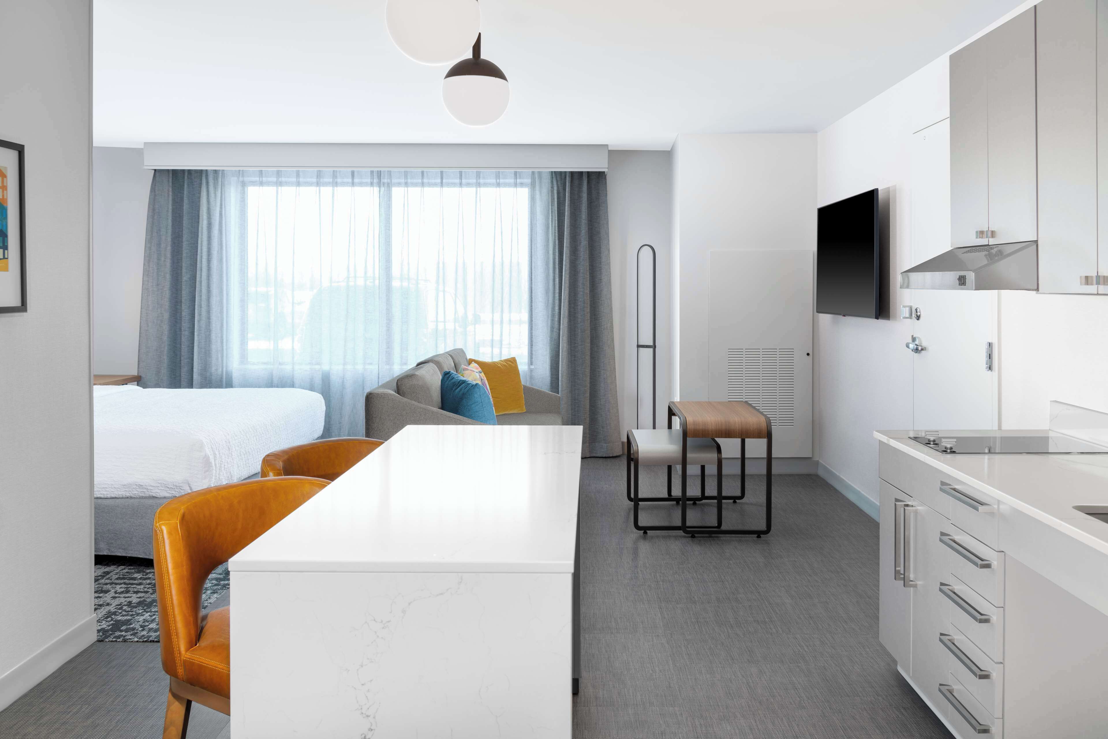 A modern hotel room with a bed, a dining area, a kitchen, and a large window with curtains.