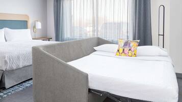Habitación de hotel con dos camas, un sillón gris, una mesita de noche con lámpara y un amplio ventanal con cortinas.