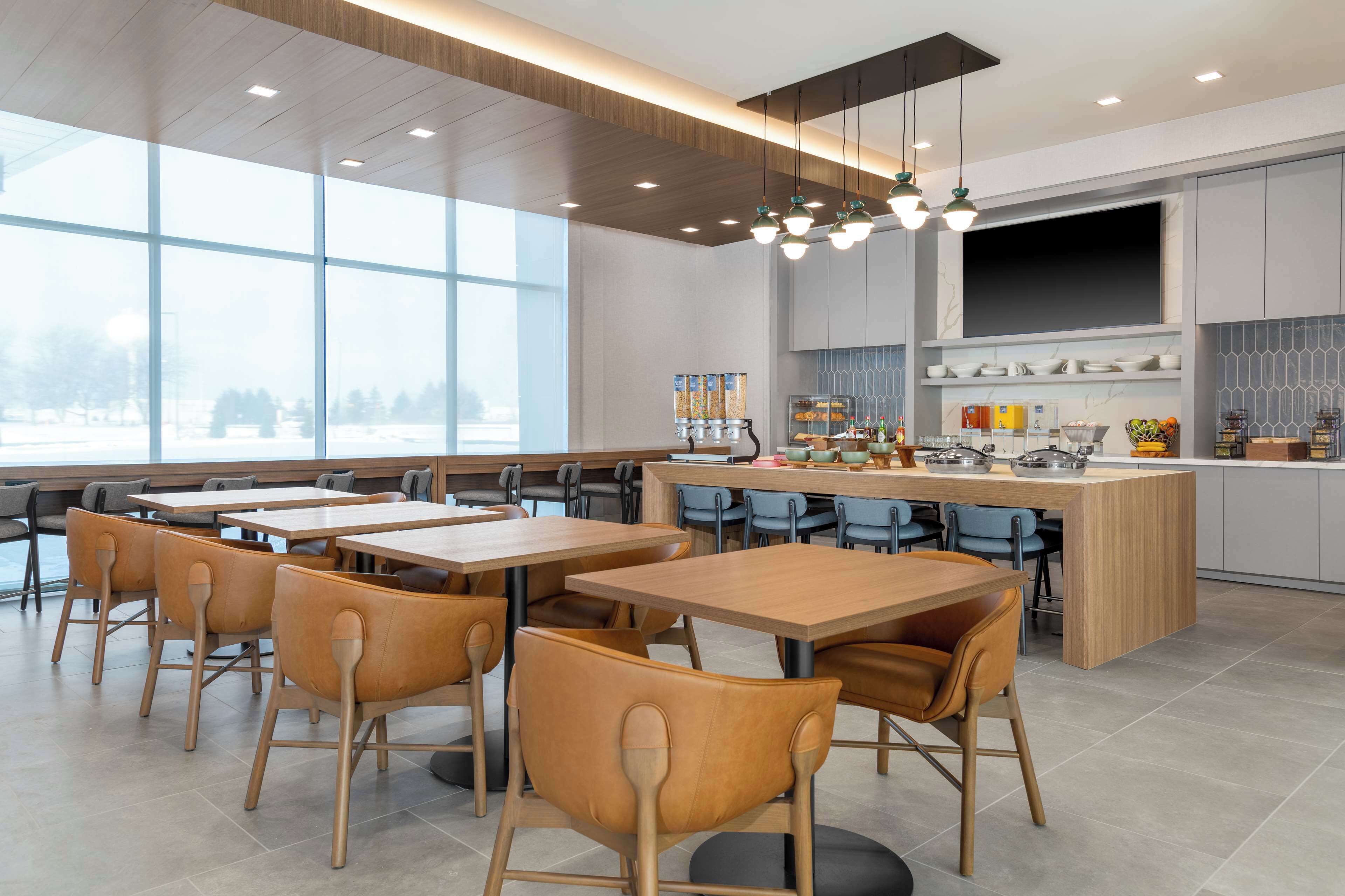A modern dining area with wooden tables and chairs, a central island with kitchen appliances, and large windows offering a view of the outdoors.