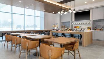 A modern dining area with wooden tables and chairs, a central island with kitchen appliances, and large windows offering a view of the outdoors.