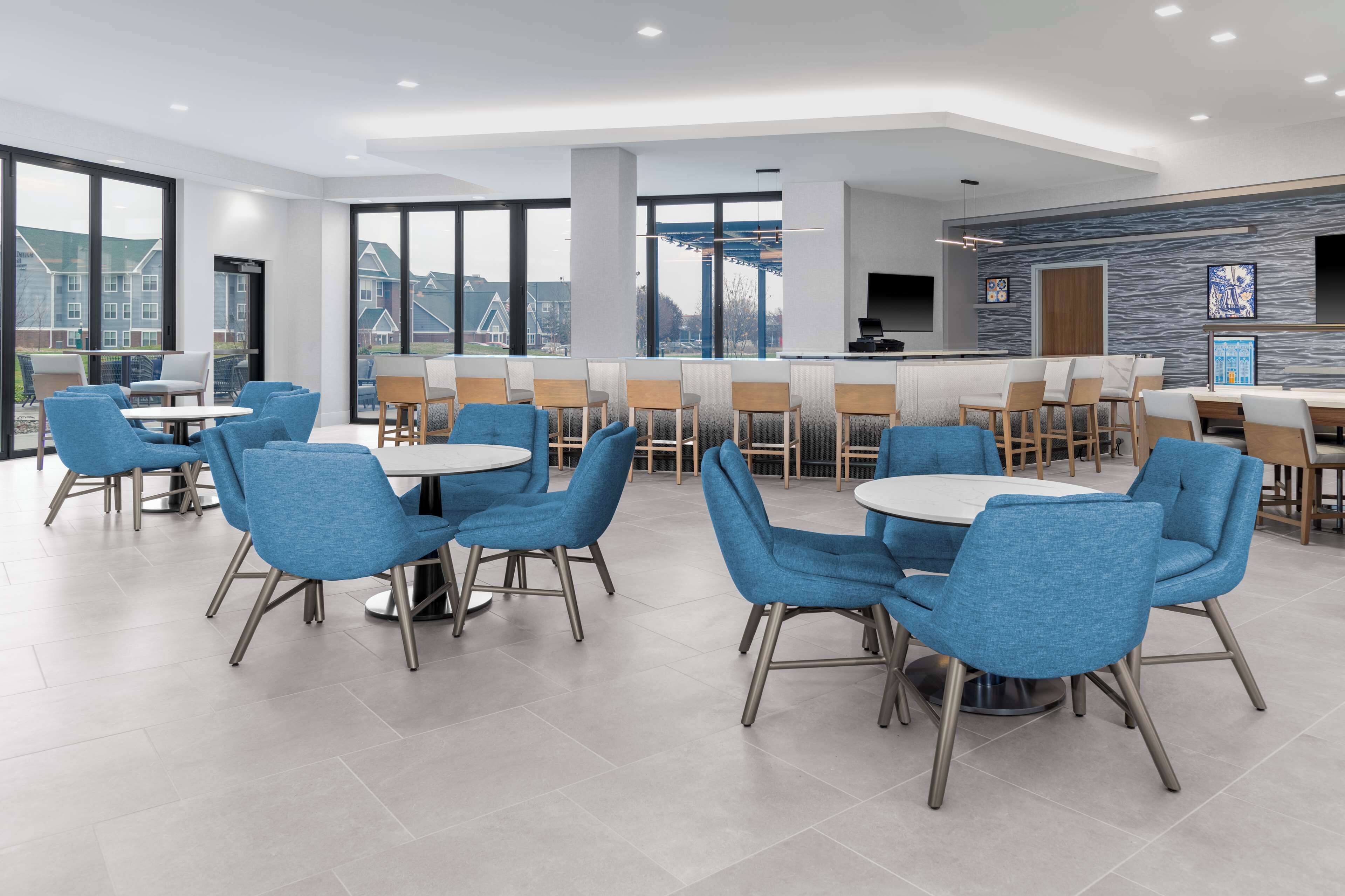 A modern dining area with blue chairs and round tables, large windows offering a view of buildings, and a bar area with a stone wall and artwork.