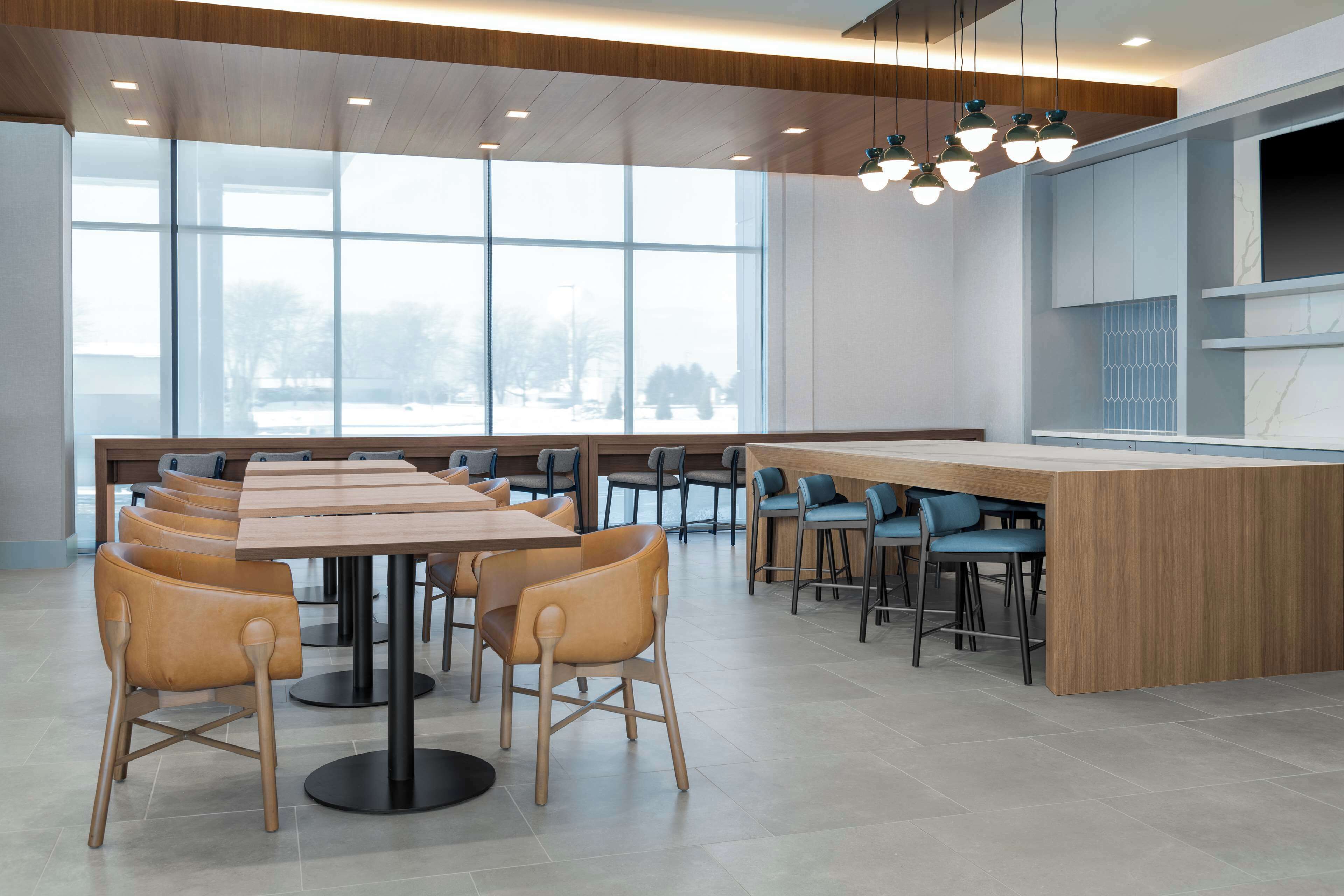 A modern dining area with a long wooden table and multiple chairs, large windows, and a kitchen area in the background.