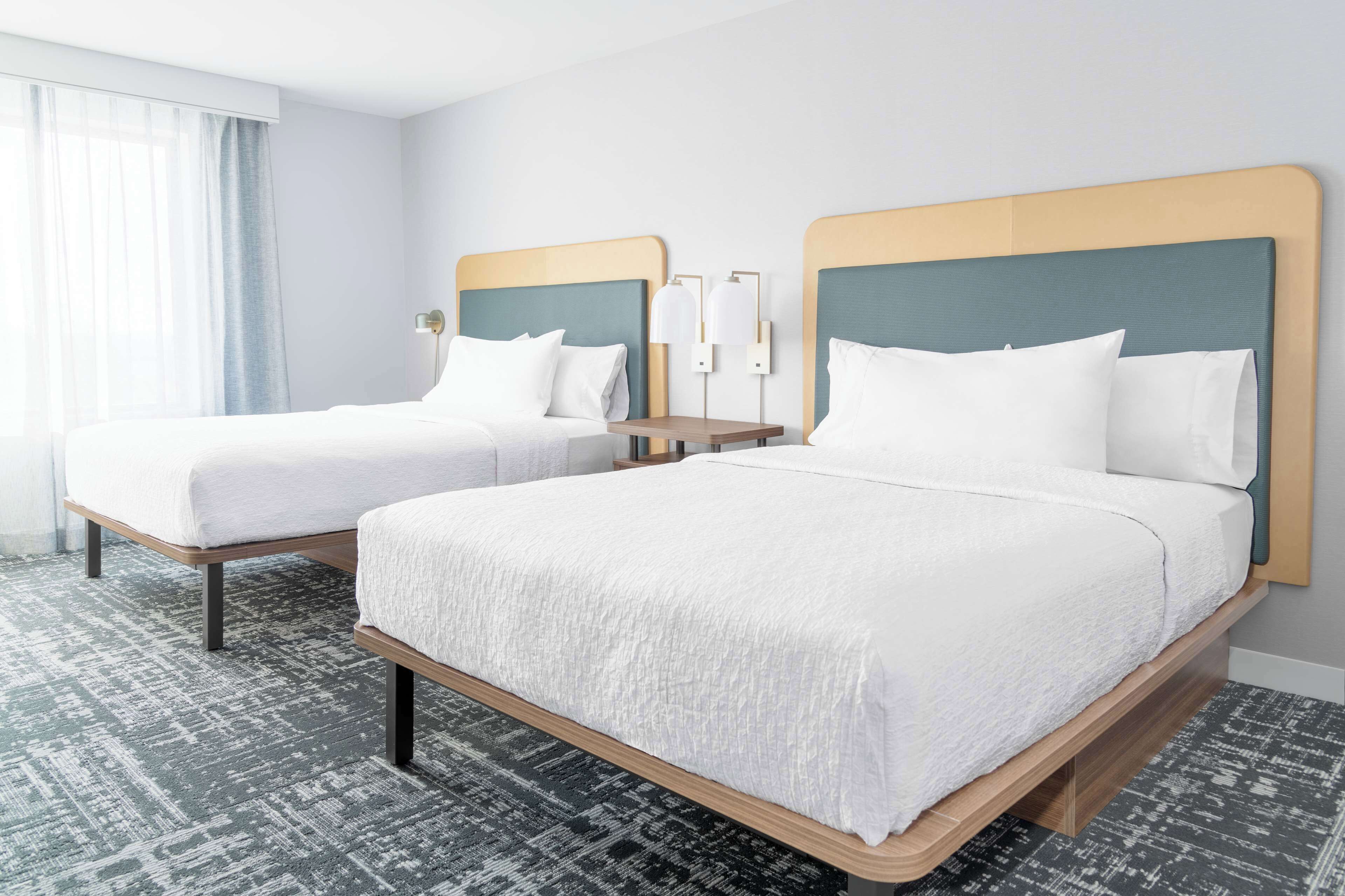 Une chambre d’hôtel avec deux lits, une grande fenêtre avec des rideaux légers, une table de chevet avec une lampe et une moquette à motifs.