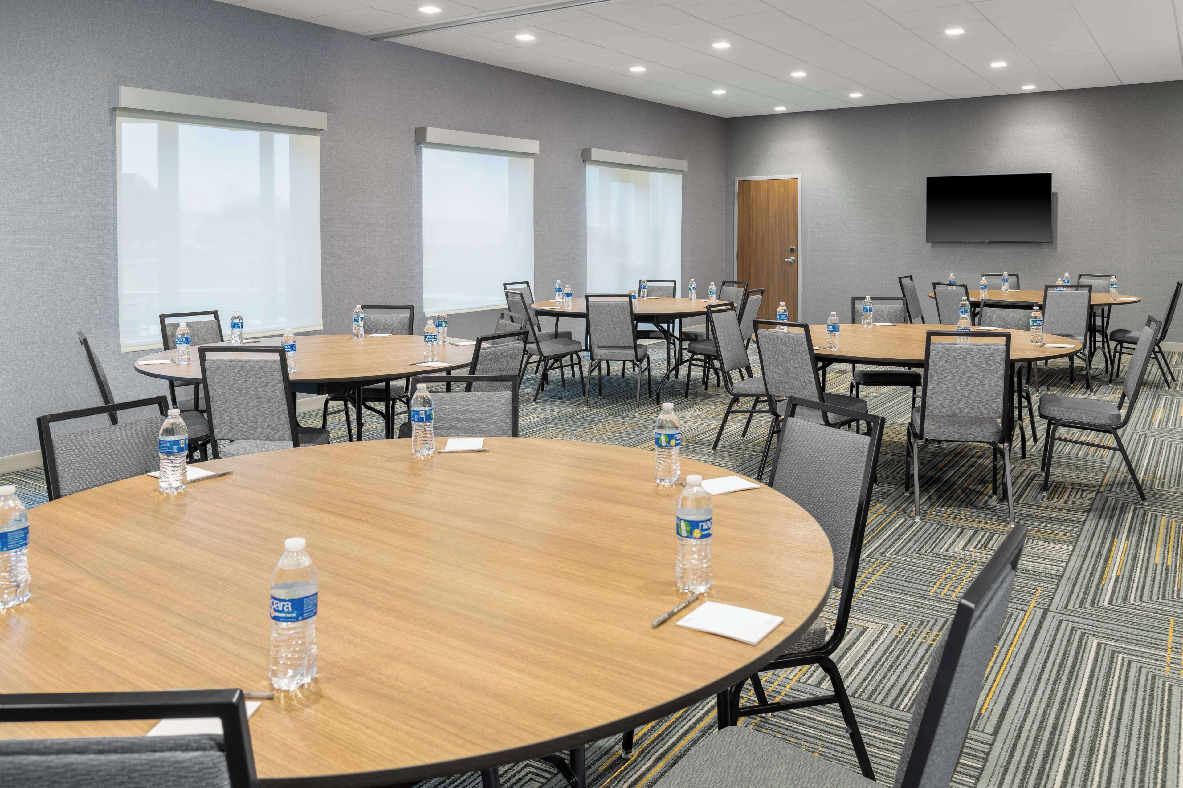 A conference room with multiple tables and chairs, equipped with water bottles, a flat-screen TV, and large windows.