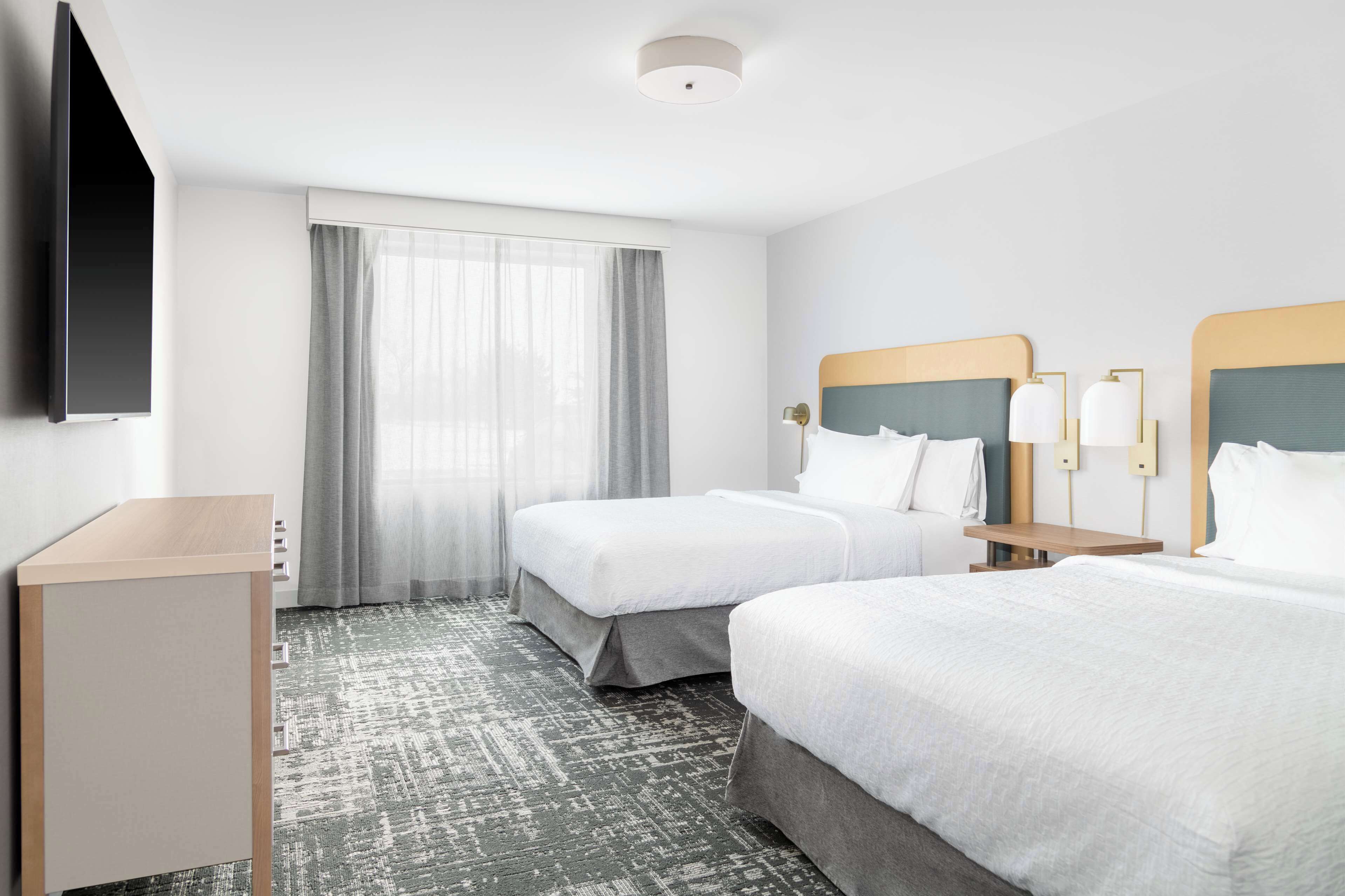 A hotel room with two beds, a TV mounted on the wall, a desk, and a large window with curtains.