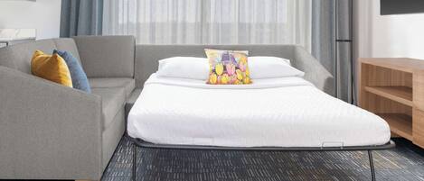 Una habitación de hotel moderna con un sofá gris, una cama blanca con un cojín colorido y una mesita de noche de madera.