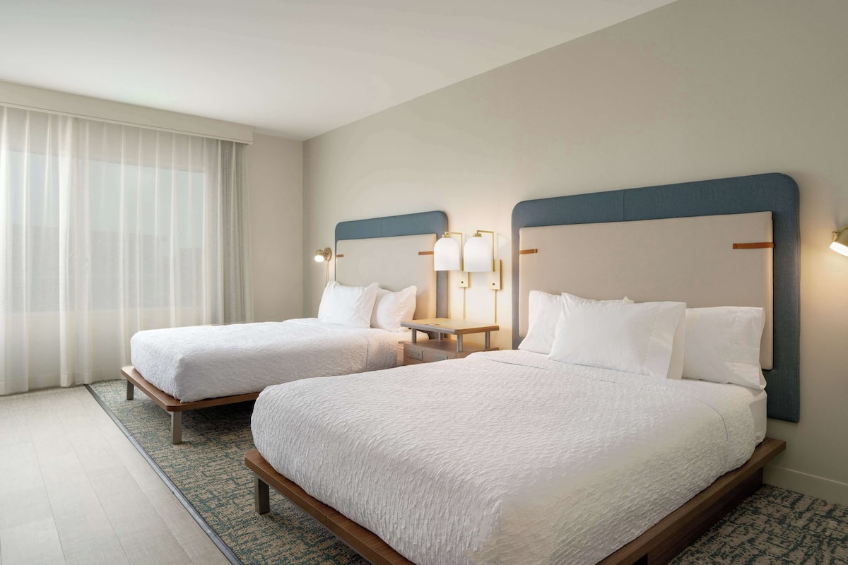 A hotel room with two beds, each with white bedding and wooden headboards. There are bedside tables with lamps and a large window with sheer curtains.