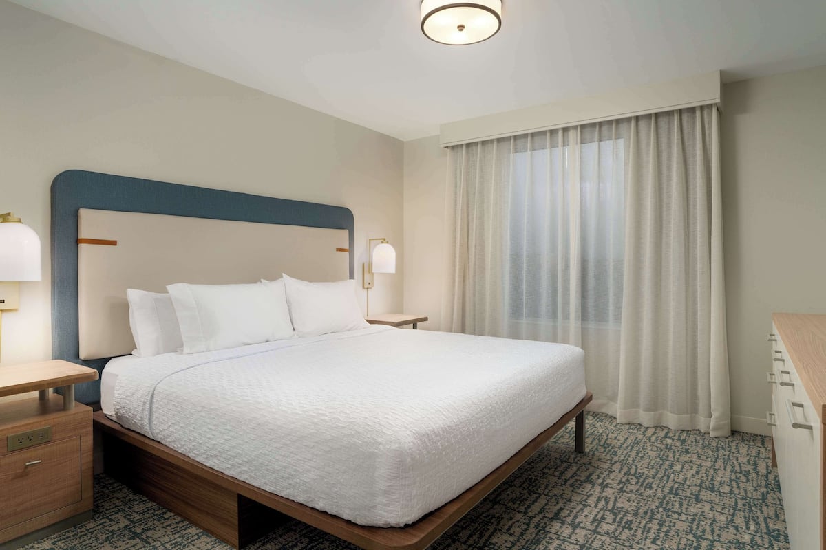 A neatly made bed with a white bedspread, a wooden headboard, a bedside table with a lamp, and a window with sheer curtains.