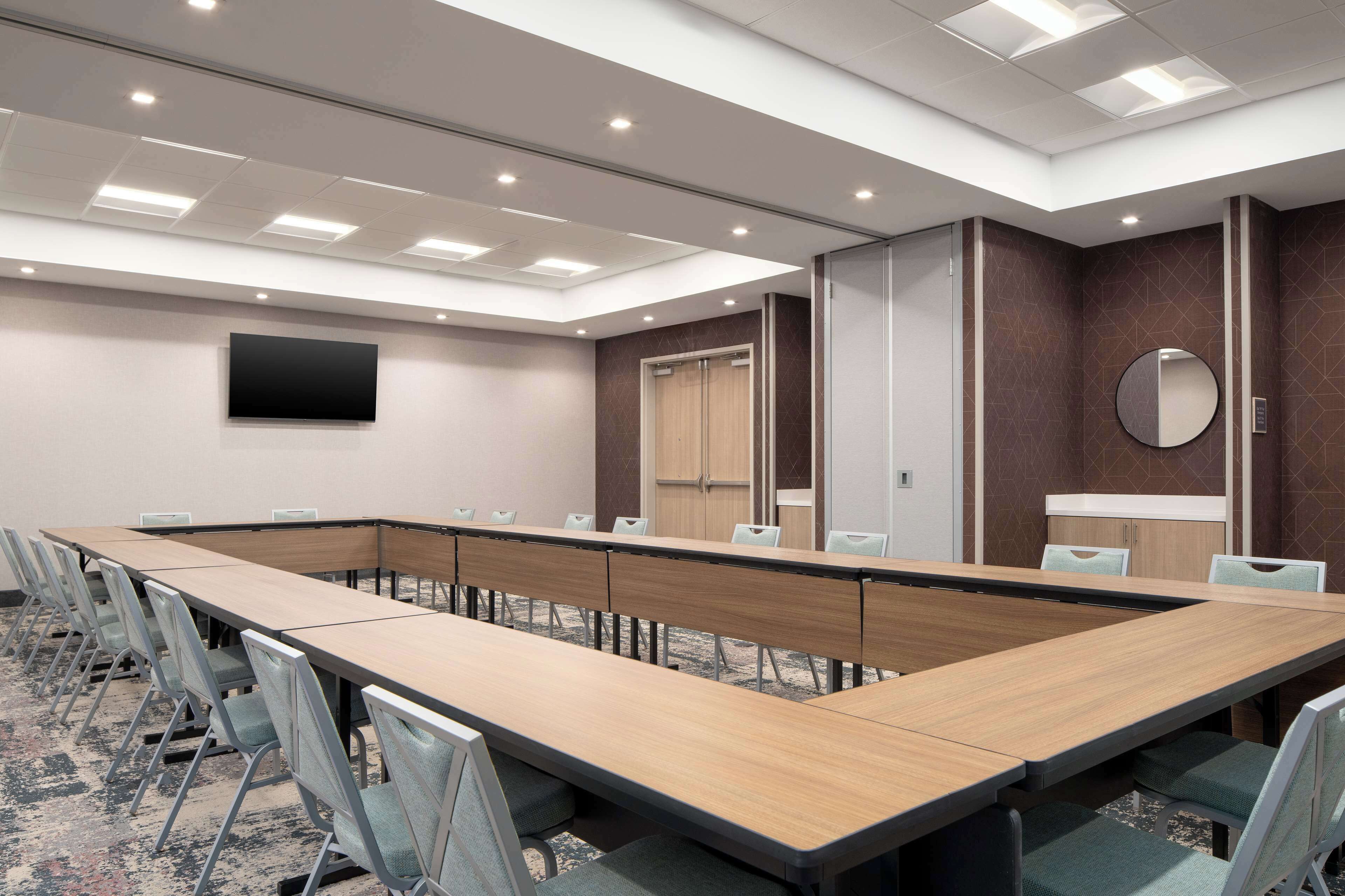 A conference room with a long table, chairs, a flat-screen TV, a mirror, and a door.