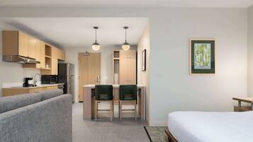 A compact hotel room with a kitchenette, a dining area with two chairs, a bed, and a framed artwork on the wall.