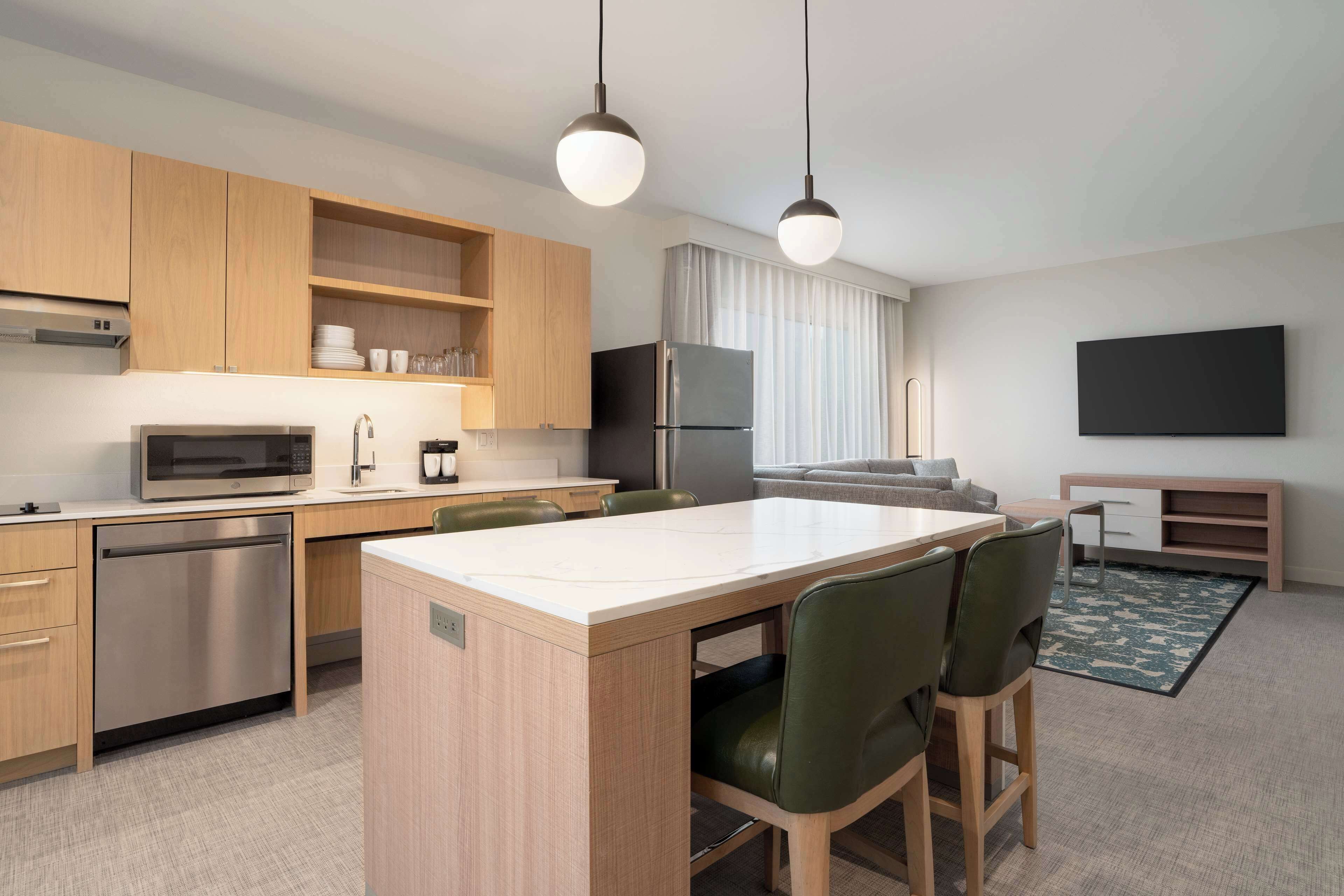 A modern kitchen with wooden cabinets, a stainless steel refrigerator, a microwave, and a dishwasher. There is a dining area with a table and chairs.