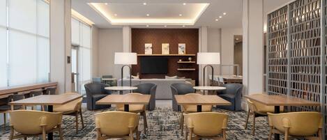 A modern hotel lobby with a seating area, tables, and a television screen.