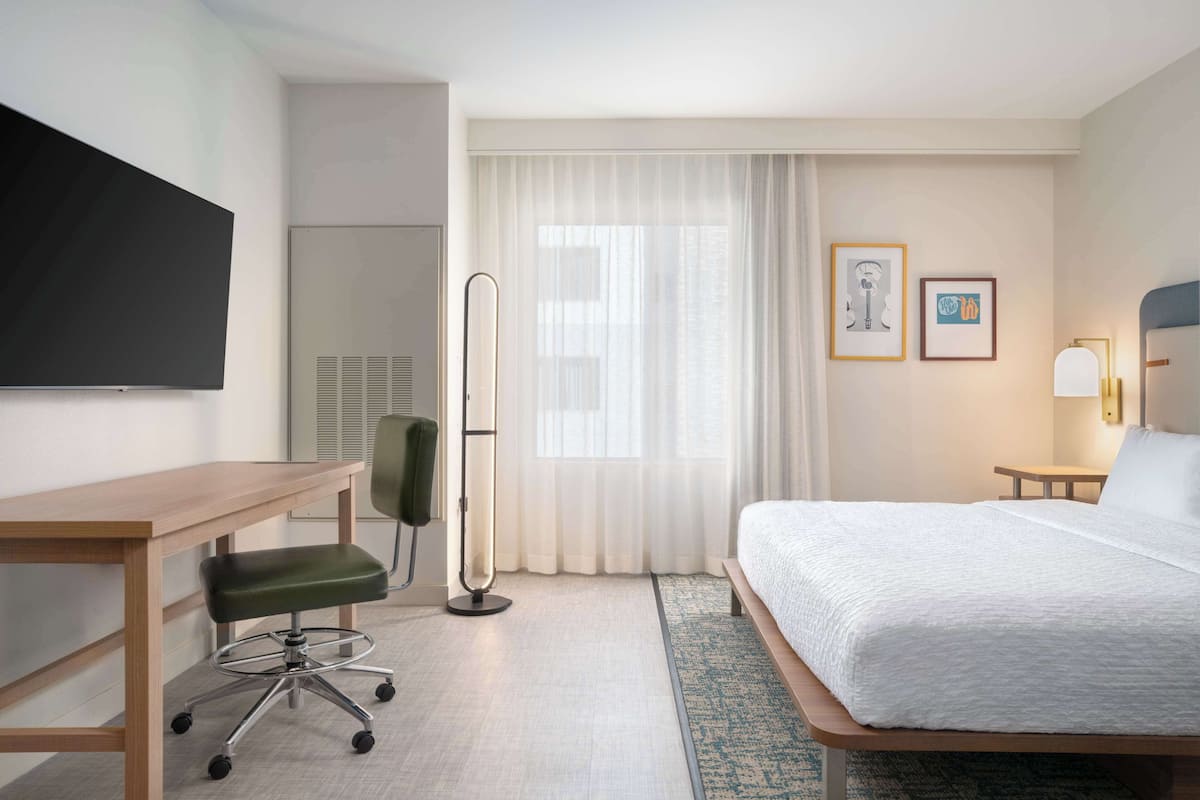 A hotel room with a bed, a desk, a chair, a TV, and a window with curtains.