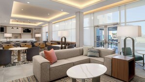 Eine moderne Hotelhalle mit einer Couch, einem Couchtisch und Lampen. Im Hintergrund sind Esstische und Stühle zu sehen.