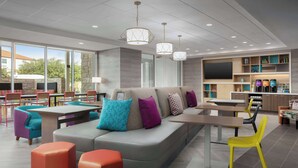 A modern lounge area with a sofa, ottomans, and chairs, a dining area with tables, and a kitchen with appliances and shelves. - Home2 Suites By Hilton Laredo North (Laredo)