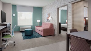 A hotel room with a desk, a pink sofa, a blue ottoman, a lamp, and a large window. - Home2 Suites By Hilton Laredo North (Laredo)