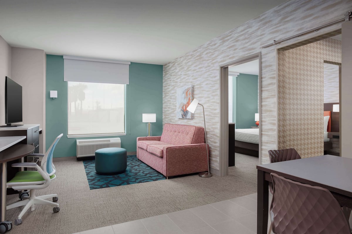 A hotel room with a desk, a pink sofa, a blue ottoman, a lamp, and a large window.
