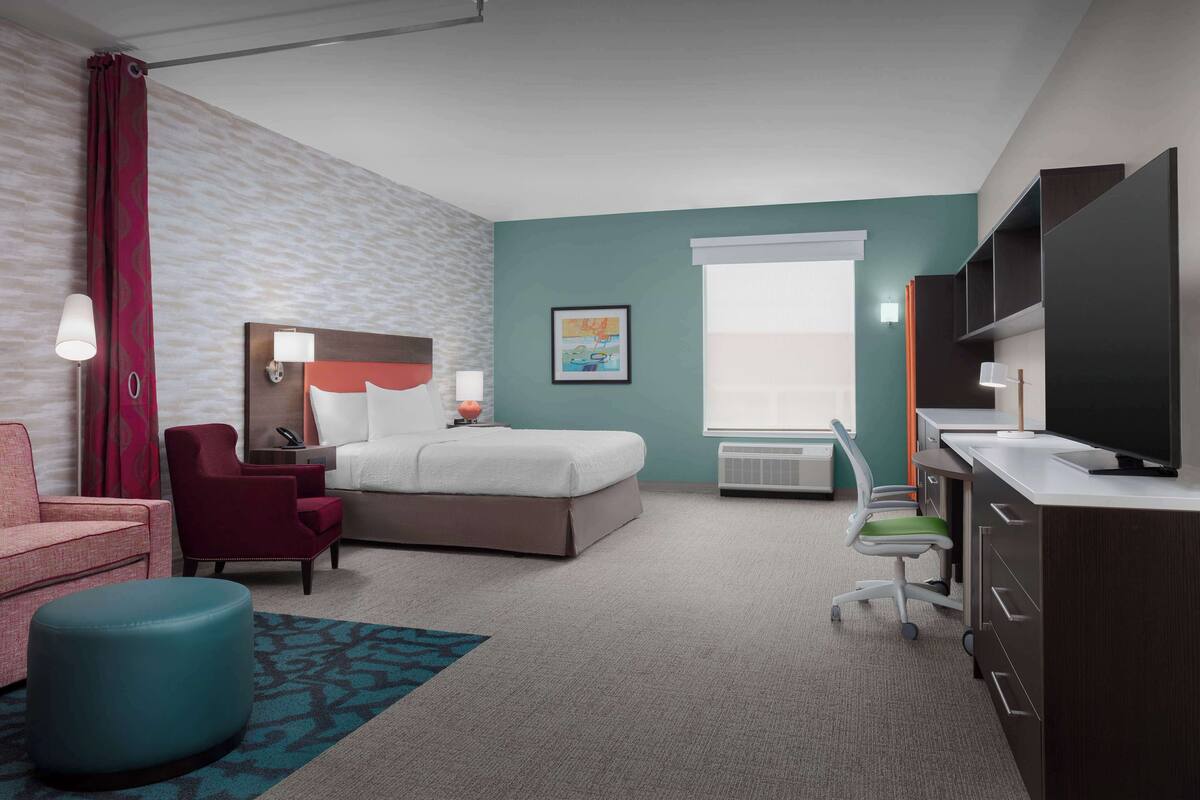 A hotel room with a bed, a desk with a TV, a chair, a desk lamp, a wall-mounted shelf, and a window.