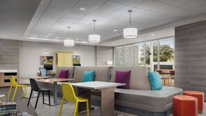 A modern interior with a dining area, a sofa with colorful cushions, and a large window providing natural light. - Home2 Suites By Hilton Laredo North (Laredo)