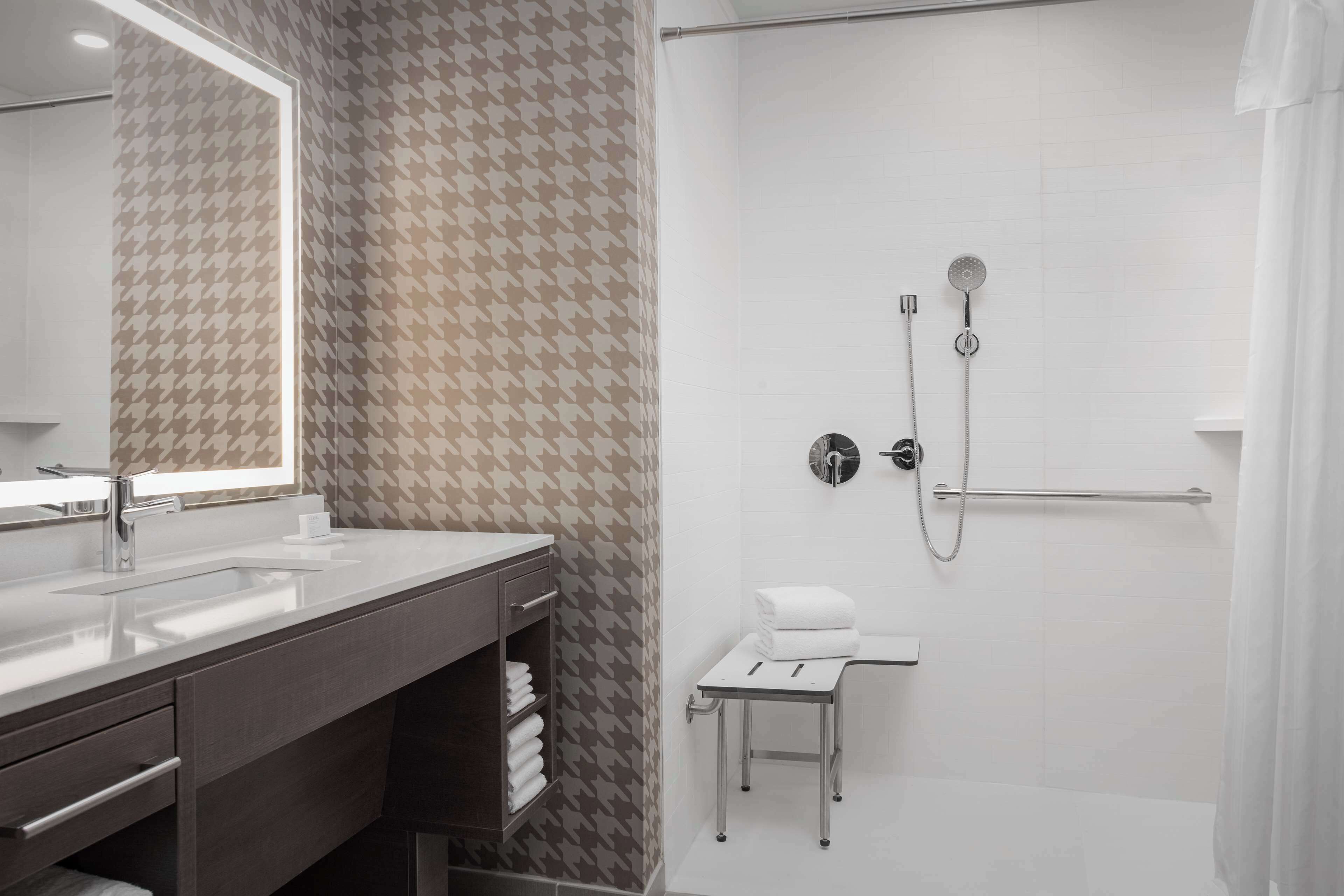 A modern bathroom with a large mirror, a sink, a shower with a handheld showerhead, and a small stool.