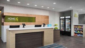 A hotel reception desk with a 'HOME 2' sign, a digital phone, and a 'Hilton' sign. There are two refrigerators labeled 'COLD' and 'COLDER' with snacks and beverages inside. - Home2 Suites By Hilton Laredo North (Laredo)